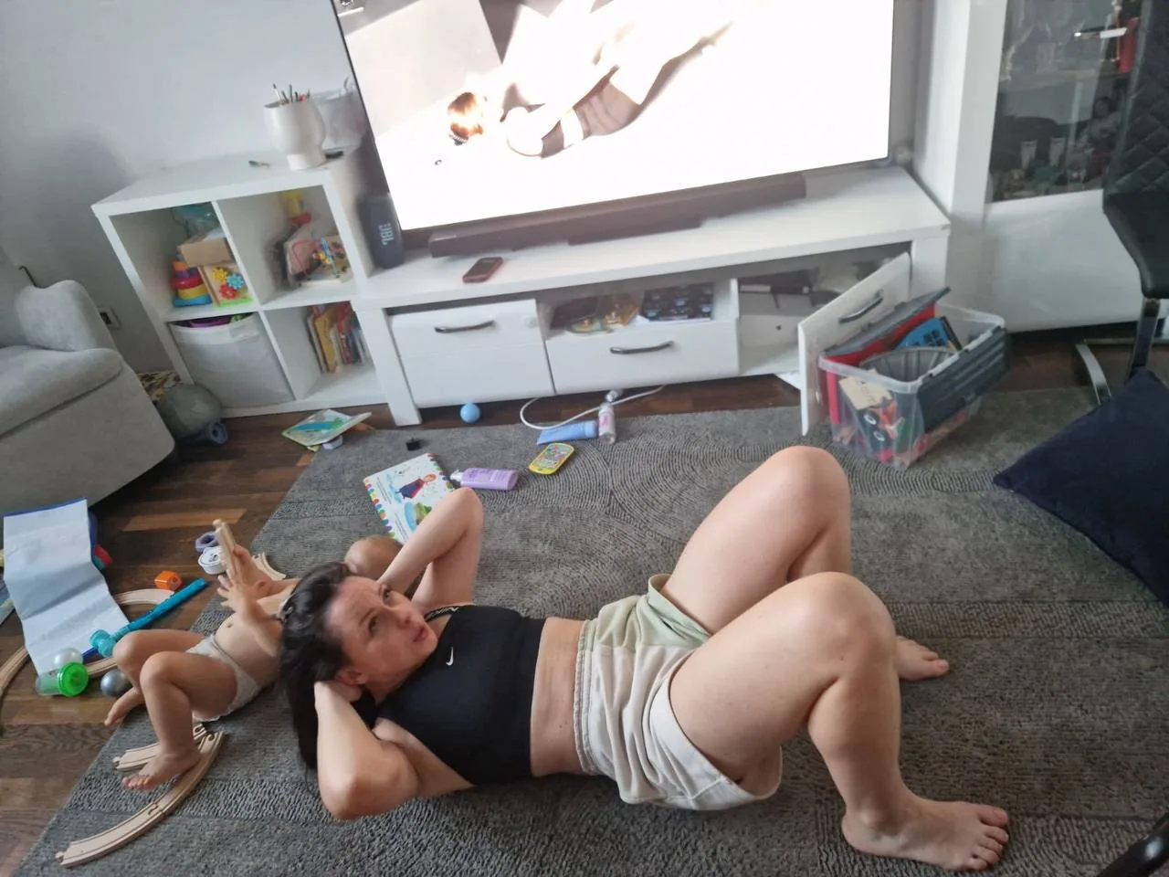 Жінка робить вправи для преса, лежачи на килимі у вітальні з дитячими іграшками поруч