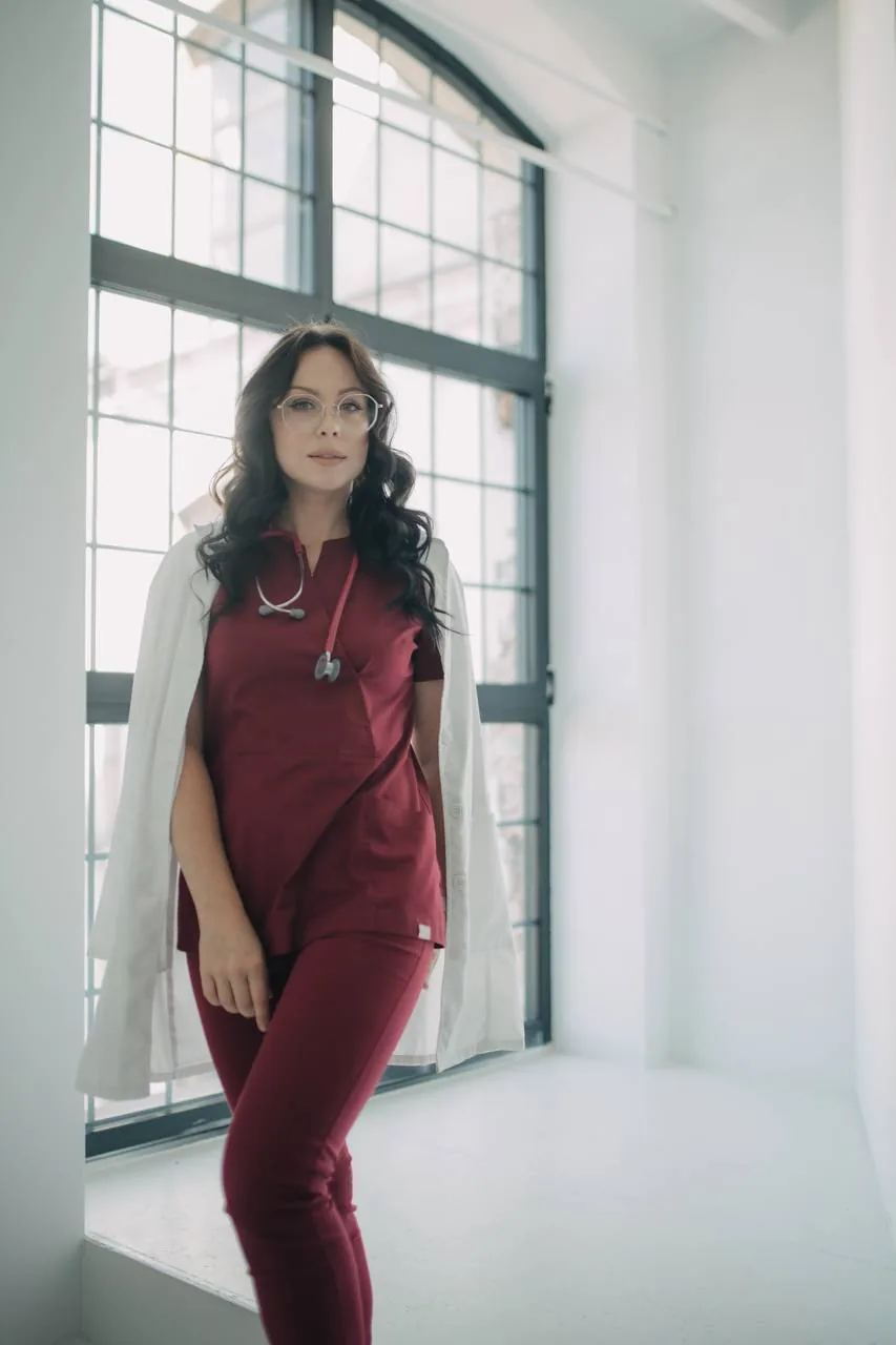 Лікарка у медичному одязі та білому халаті стоїть біля великого вікна